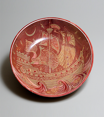 Maroon plate with a yellowy orange medieval ship decorated on the base. It has sales and portholes and there are decorations of waves below it and birds around it as if it is out to sea