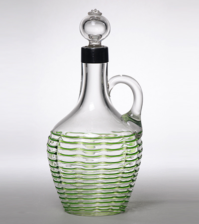 Glass jug with bottle stopper in the neck. It has a tall neck with a handle to the right, and a wide curved base decorated with translucent green lines