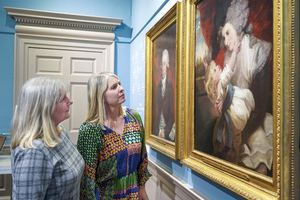 Two women stand in an art gallery, looking closely at two framed paintings. The room has light blue walls and a decorative door. The paintings appear to be classical portraits with figures in historical attire.