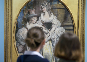 Two people stand in front of an ornate framed painting depicting two women in historical attire with hats, against a blurred background.