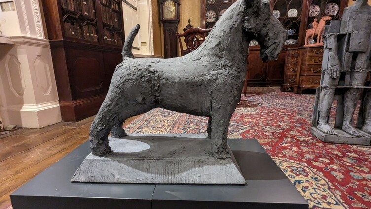 A rough-textured sculpture of a dog stands on a black platform in a room with antique furniture, bookshelves, a grandfather clock, and ornate rugs.