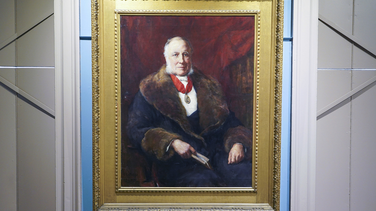 A framed portrait of an older man wearing a fur coat and a medal around his neck. He holds a book and sits against a dark red background. The painting is displayed on a blue wall.