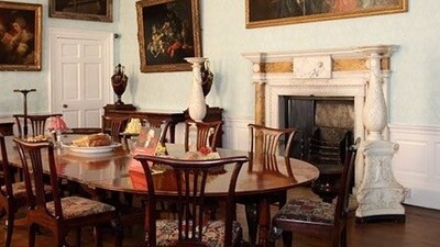 Elegant dining room with a polished wooden table, ornate chairs, a fireplace with white mantel, large vases, chandelier, and classical paintings on pale blue walls. The room has high ceilings and wooden floors.