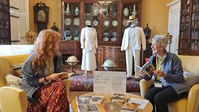 Two women sit and read books in a museum room with antique furniture and glass cabinets. Behind them are two mannequins dressed in vintage clothing and a chandelier hanging from the ceiling. Books are spread on the table between them.
