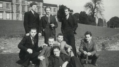 A black-and-white photo of eight men in suits posing together on grass outside a large historic building, with trees and a stone wall in the background. The caption reads: “A group of ex-soldiers in their Sunday best outside Cannon Hall.”.