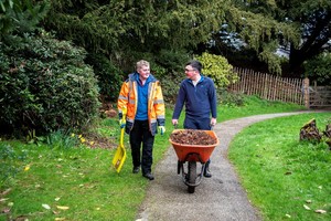 Two volunteers walking together, one pushing wood chipping in a wheel barrow and the other carrying a shovel.