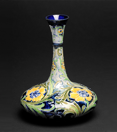 Vase with a tall thin neck and wide squat base. It is decorated with yellow flowers and green leaves with blue in the background and inside the neck