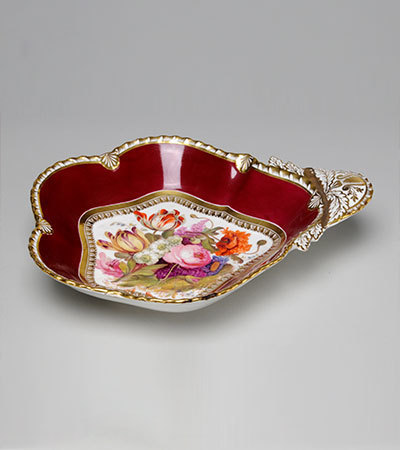 Small porcelain dish with low sides and a roughly circular base. It has white and brown edging, and a maroon colour on the inside of the sides. The base is painted with different coloured flowers, it has a handle on the lright hand side.