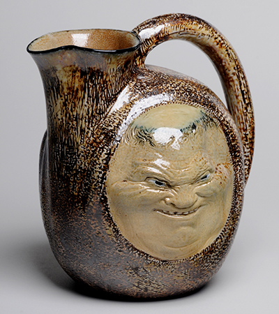 Brown jug with a round body and short spout on the left hand side. The handle starts at the spout and curves down the left body. A round light brown face with a creepy grin is decorated on the side.