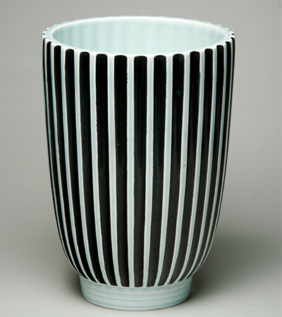 Tall vase which does not taper in much towards the base. It is white and decorated with vertical black stripes.