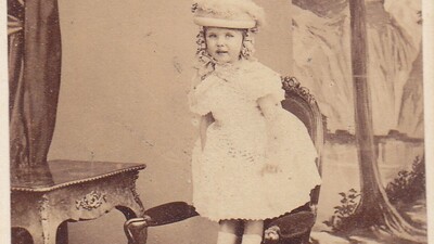 A young girl in a light dress and hat stands on a ornate chair beside a table, with a painted backdrop of a tree and mountains. Handwritten text appears at the bottom.