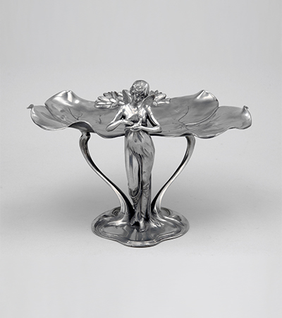 Small silver metal sculpture of a woman standing with a bird in her hands looking down. Behind her, the visiting card tray stretches out in the form of a large leaf.