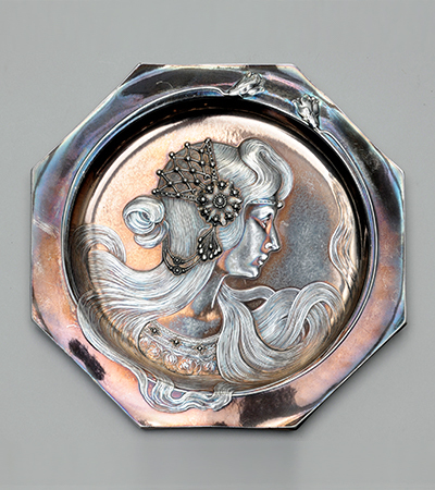 Octagonal metal plate with the side profile of a woman looking to the right. She wears an ornate headdress and her hair is long and wavy