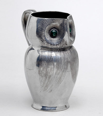 Small silver pot in the rough shape of an owl, with the base as the body. The head of the owl has two eyes looking forward and the jug tapers to a point where the beak is to pour the liquid in it. There is a silver handle at the back of the jug.