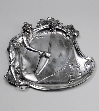 Silver tray decorated with a woman on the left filling up a long thin jar with water. The edges of the tray are decorated with silver foliage.