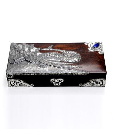 Wooden rectangular shaped box with the opening on the long side. Decorated with silver metal geometric designs and swirls and one blue gemstone in the top right corner.