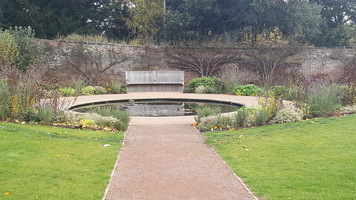 Circular pond in walled garden, surrounded by plants. There is a bench to sit on.