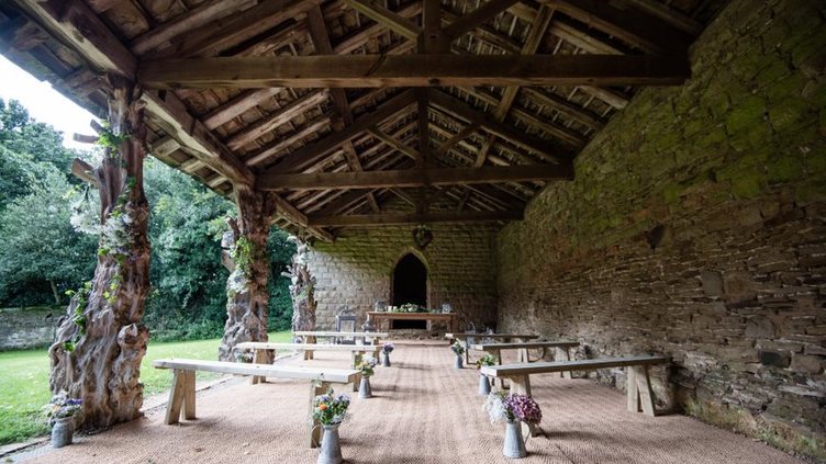 A long space with a stone wall on one side and ornate colonnades on the other. Benches are set out with flowers in pots at the end of them.