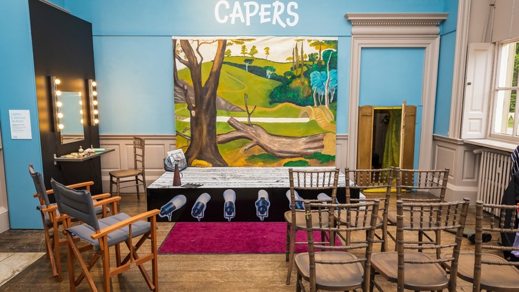 A room with wooden chairs and a makeup vanity on the left. A stage with various hats under spotlights is in front of a large painting of a park scene. "COSTUME CAPERS" is written on the blue wall above the painting. A window is on the right.
