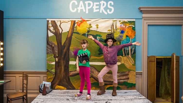 Two children stand on a stage in front of a painted forest backdrop. One child wears pink leggings and a black shirt, the other wears pink pants, a pink hoodie, and a black hat. Above them, the sign reads "Costume Capers.