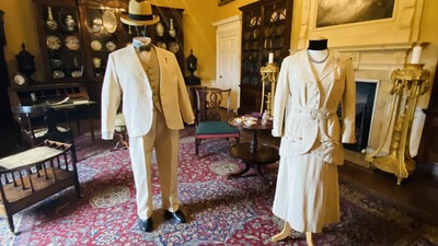 Two mannequins dressed in vintage white formal attire stand in a luxurious room with ornate furniture. The room features a large, colorful area rug, elegant wooden cabinets, and framed paintings on yellow walls under a chandelier.