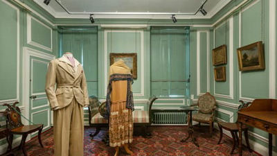 A room with light green walls and a red patterned carpet displays two outfits on mannequins: a tan suit and a brown dress with a shawl. The room also contains paintings, vintage chairs, and a wooden desk.