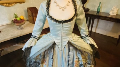A mannequin displays an elaborate historical dress with lace details and floral embroidery. The gown features a light blue satin fabric with a black lace trim,