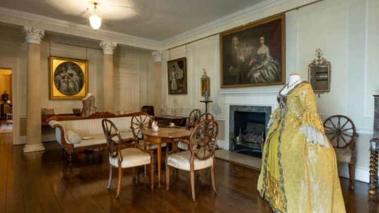 A vintage room featuring a yellow period dress on a mannequin, a wooden dining table with chairs, classic sofas, and framed paintings on walls. The room has wooden floors, columns, and a fireplace, exuding a historical ambiance.