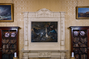 A lavish room features an ornate fireplace with an intricate stone mantle. Above it hangs a dramatic painting of a mythological scene. Flanking the fireplace are two tall wooden bookshelves with glass-enclosed cabinets and decorative plates.