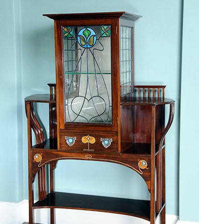 Art nouveau cabinet with wooden legs and frame and a glass door decorated with black lines in the shape of a heart and geometric shapes. There are three small stained glass panels at the top.