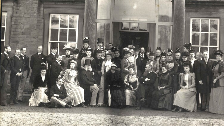 A large group of men and women in Victorian-era clothing pose together outside a grand stone building with tall columns and large windows. Some people are seated while others stand around them.