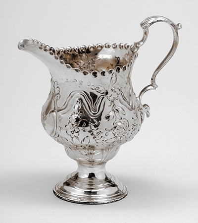 Ornately decorated silver jug with handle to the right and spout to the left. It has a round base and short stem, and ornate decorations of foliage carved into it