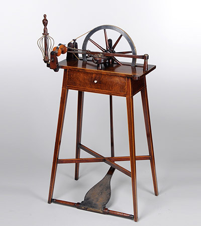Wooden spinning wheel with pedal and four legs. Only the top half of the wheel is visible with spinning apparatus to the left hand side.