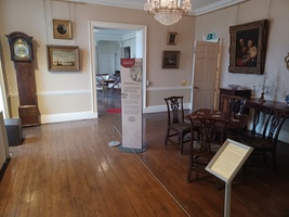 Games room. A card table to the right of the image with artworks on the wall and a Grandfather clock to the left.