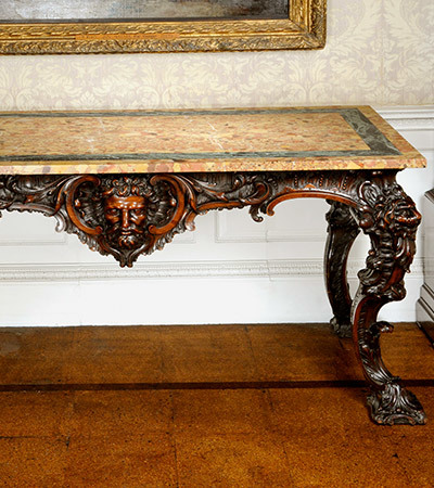 Marble table with wooden carved legs standing against a wall. The legs have faces of Greek figures carved into them.