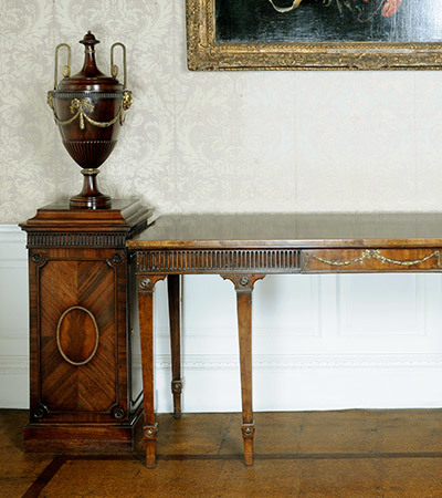 A wooden table. To the left of it is a tall thin wooden stand with an wooden urn on top of it decorated with gold designs.