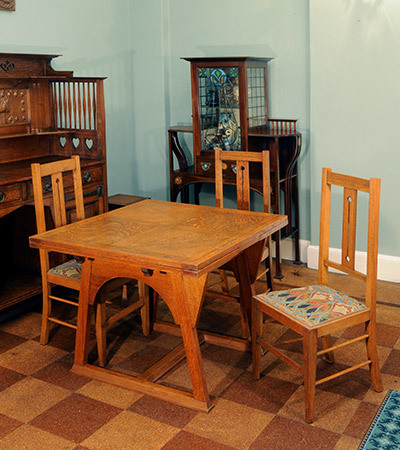 Square wooden table and three chairs around it. The wood is light coloured and the chairs have cushions with geometric decorations